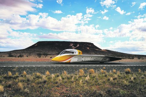 Flaches, aerodynamisch geformtes Solarfahrzeug des Teams Sonnenwagen fährt auf einer Straße durch eine karge Landschaft unter blauem, bewölktem Himmel.