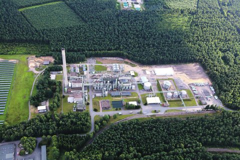 Luftaufnahme eines von Wald umgebenen Industrieareals mit Produktionsanlagen, Tanks, Schornstein, Lagerhallen, Straßen und angrenzenden Solarmodulen.